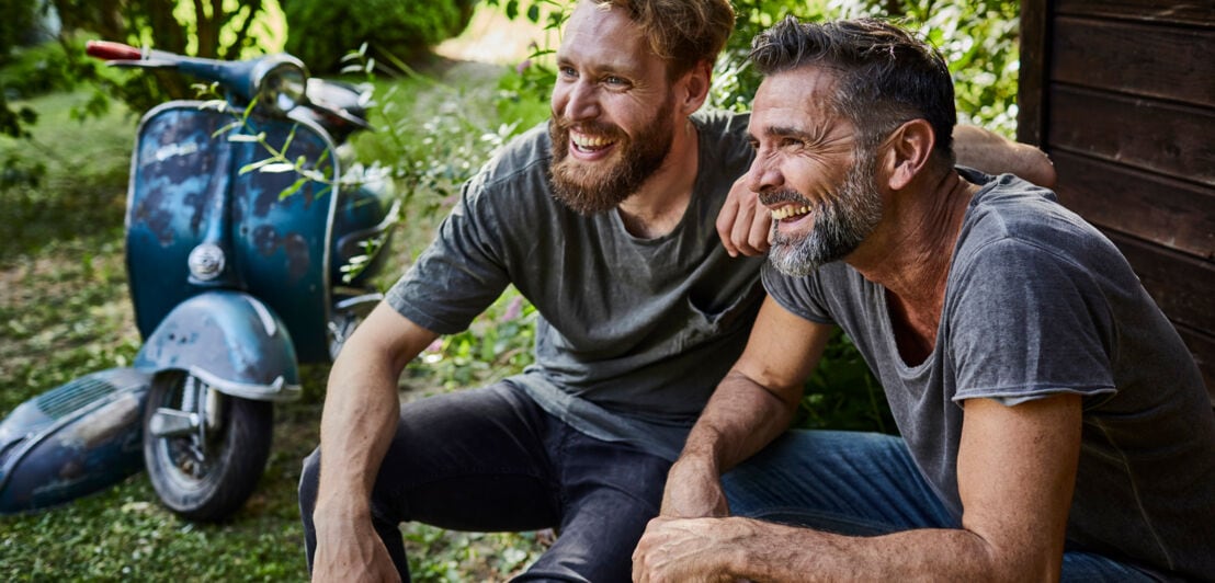 Zwei junge Männer in T-Shirts mit Vollbärten lachen zusammen im Park, im Hintergrund eine Vespa