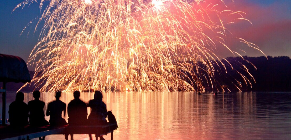Mehrere Personen sitzen auf einem kleinen Steg an einem See, über dem ein Feuerwerk stattfindet