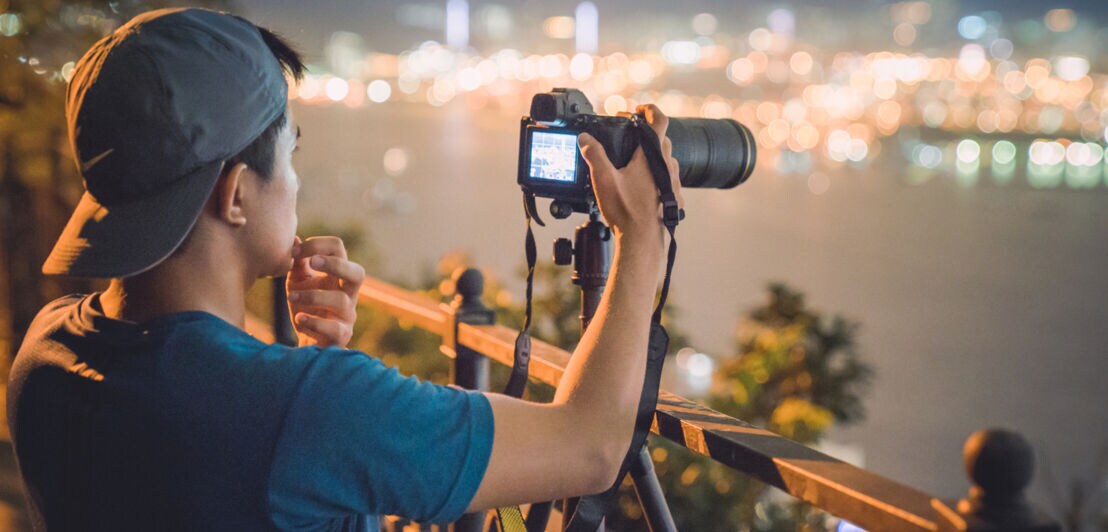 Ein Mann mit Käppi steht vor einem Geländer über einer Stadt und stellt etwas an einem Fotoapparat auf einem Stativ ein