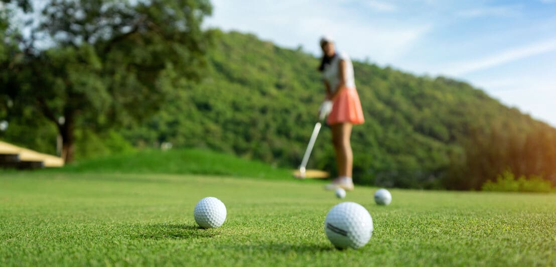 Im Vordergrund Golfbälle auf dem Rasen, im Hintergrund eine Frau mit Golfschläger