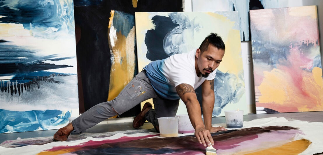 Ein kniender Mann malt ein großformatiges, abstraktes Bild auf dem Boden