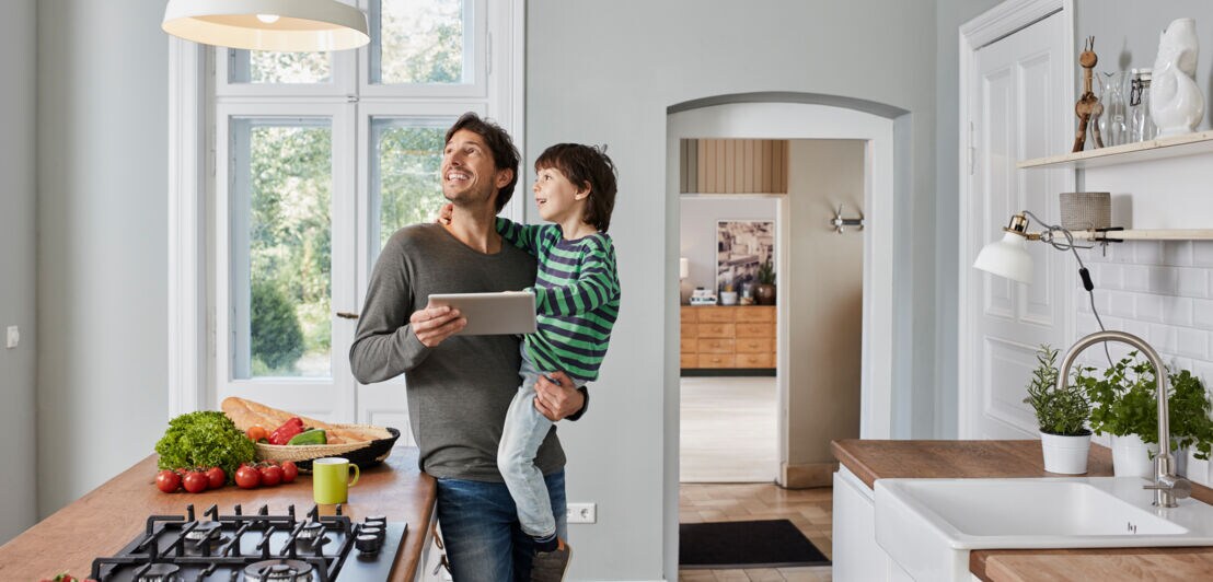 Ein Mann mit Kind auf dem Arm und Tablet in der Hand steht staunend in der Küche