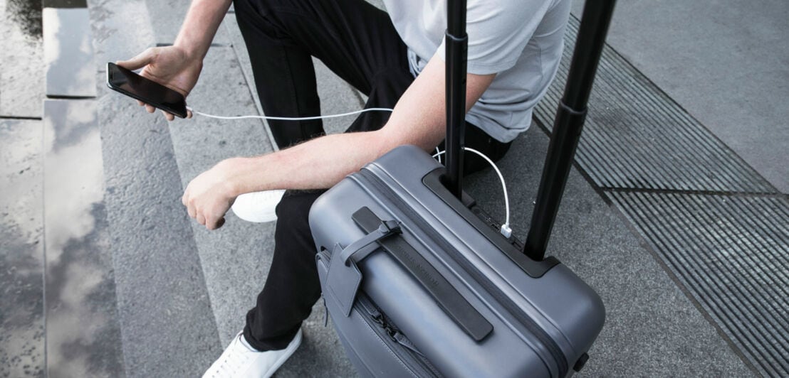 Ein Koffer steht neben einem Mann der ein Handy hält das über USB am Koffer angeschlossen ist