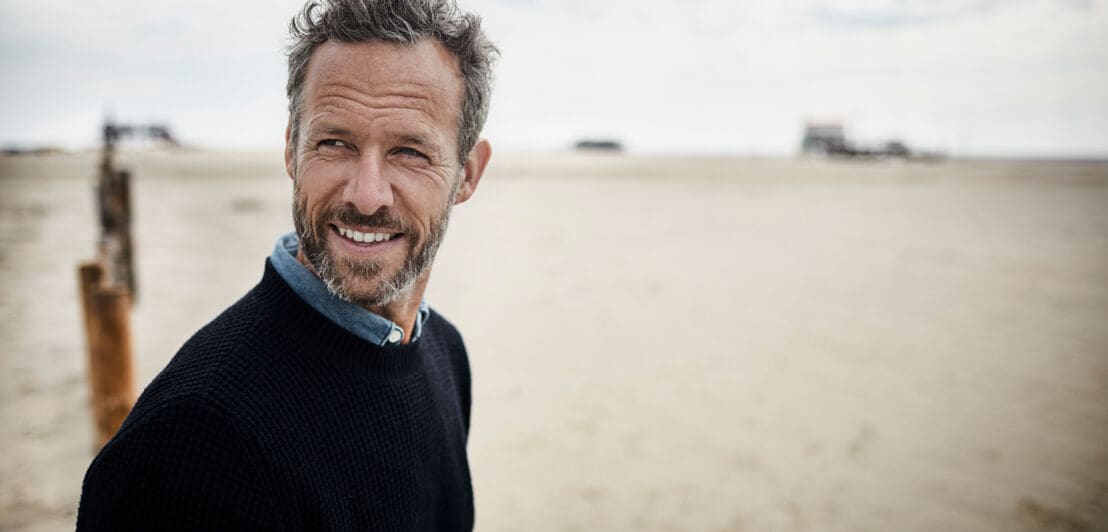 Ein Mann mittleren Alters mit Bart steht am Strand und lächelt