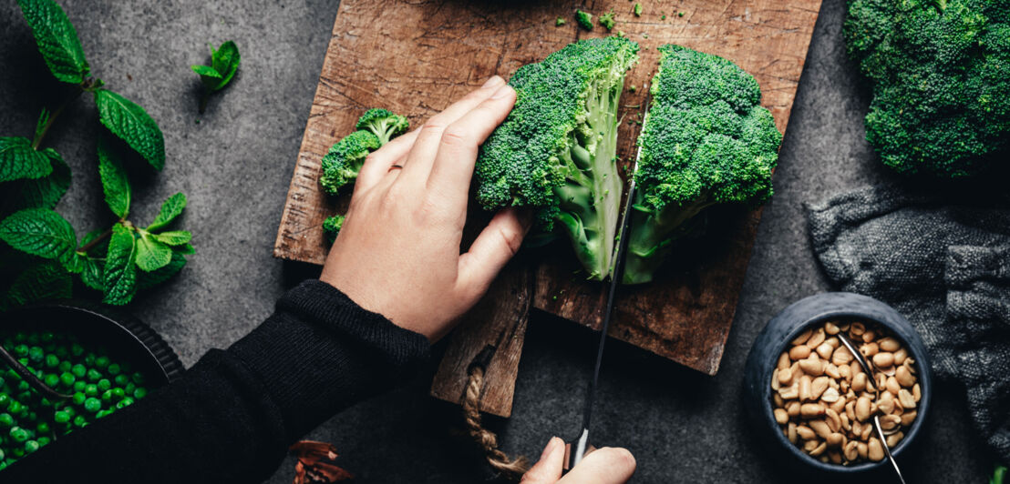 Aufsicht einer Küchenszene mit zwei Händen, die Brokkoli auf Brett schneiden