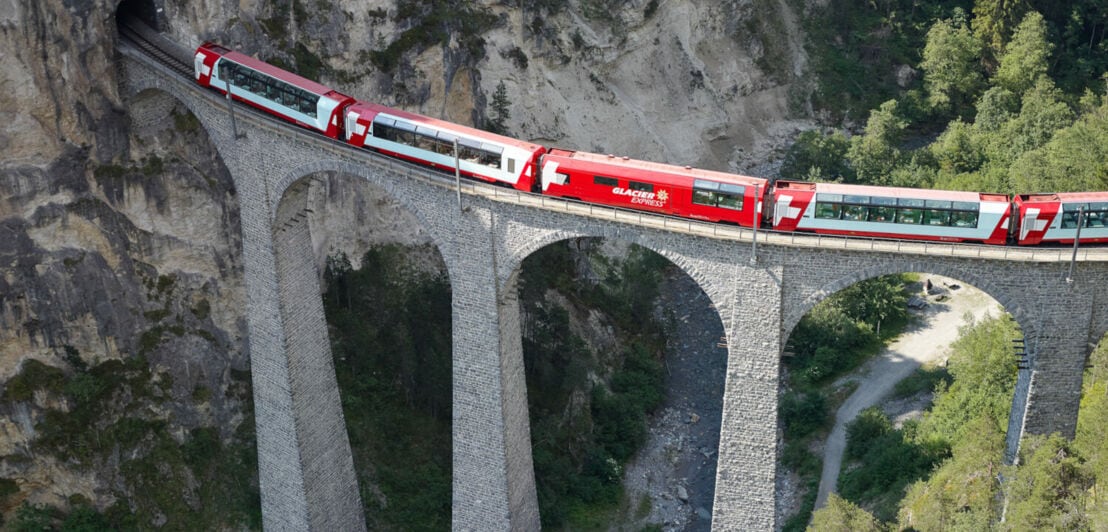 Vogelperspektive eines roten Zuges, der auf einem Viadukt durch eine Berglandschaft fährt
