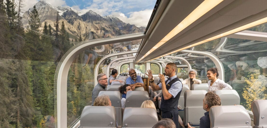 Passagiere in einem Zugabteil mit rundum Verglasung mit Panoramasicht auf Berglandschaft