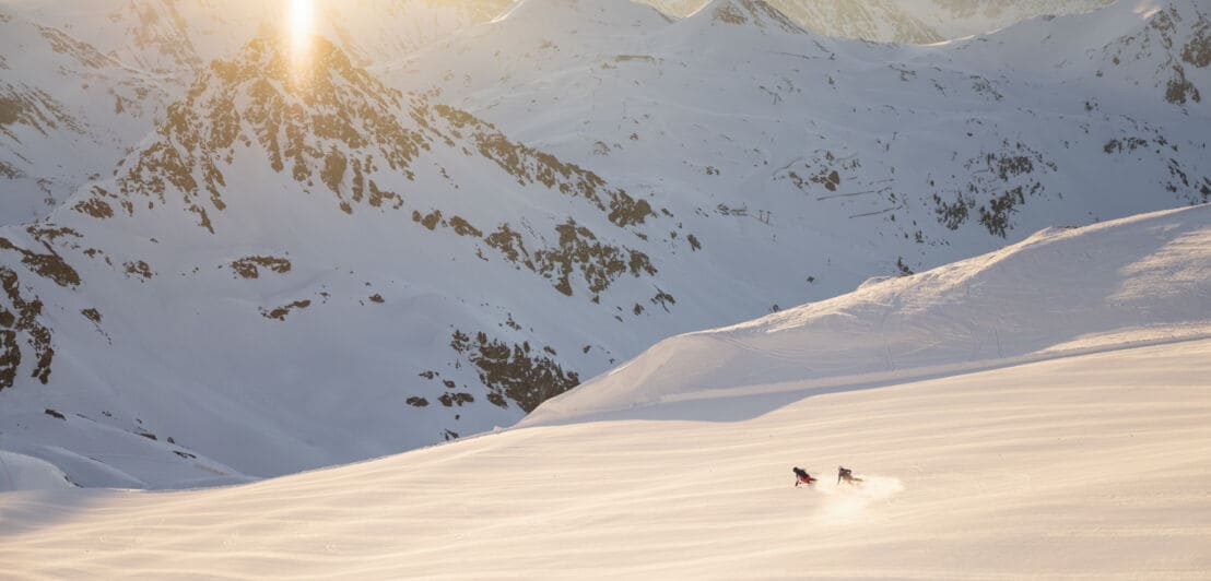 Zwei Skifahrer:innen fahren eine Piste hinab, im Hintergrund eine Bergkette