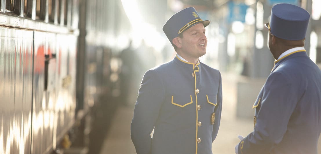 Ein Zug steht im Bahnhof, daneben zwei männliche Zugbegleiter in blauen Uniformen auf dem Bahnsteig