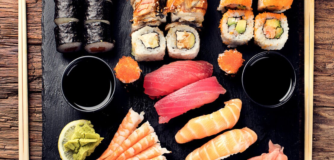 Aufsicht unterschiedlicher Sushi-Sorten mit Beilagen auf schwarzem Tablett
