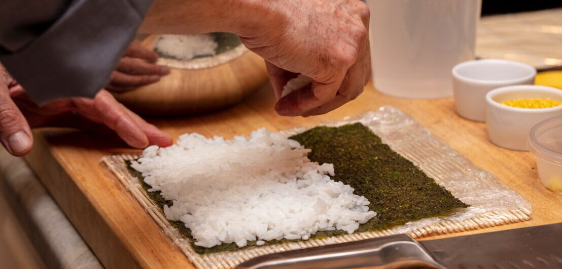Eine Lage Reis wird von einer Hand auf einem Algenpapier auf einem Brett angerichtet