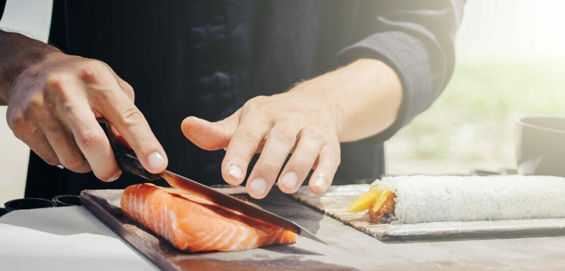 Zwei Hände mit Messer bearbeiten ein Stück Lachs auf einem Brett, daneben eine Reisrolle