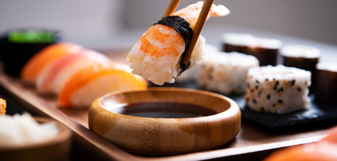 Ein Stück Nigiri-Sushi mit Garnele wird von zwei Essstäbchen über einer Holzschale mit Sojasauce gehalten, im Hintergrund weitere Sushi-Stücke auf Tablett