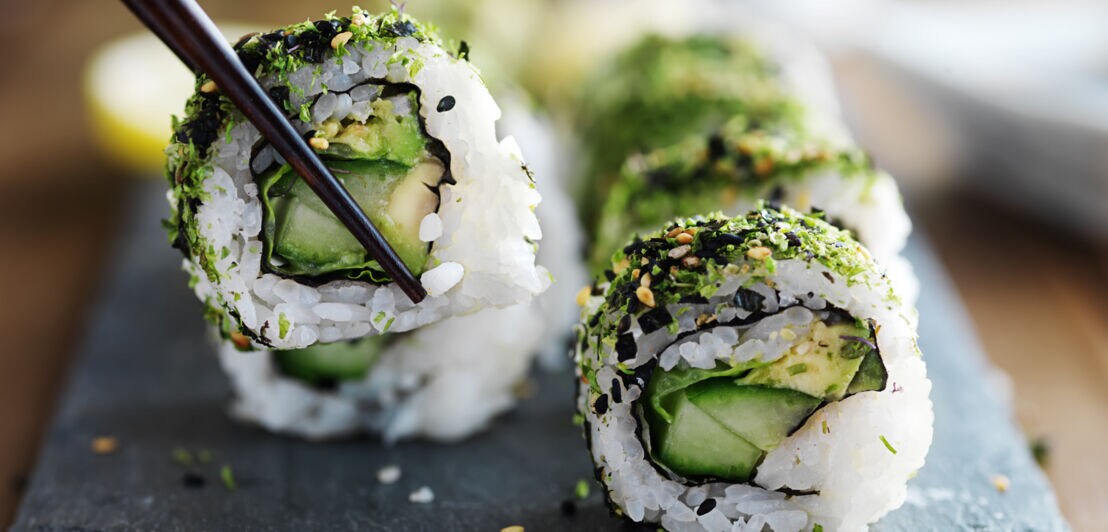 Nahaufnahme einer geschnittenen Sushirolle mit vegetarischer Füllung und grünem Topping auf Schieferplatte, ein Stück wird von Essstäbchen gegriffen