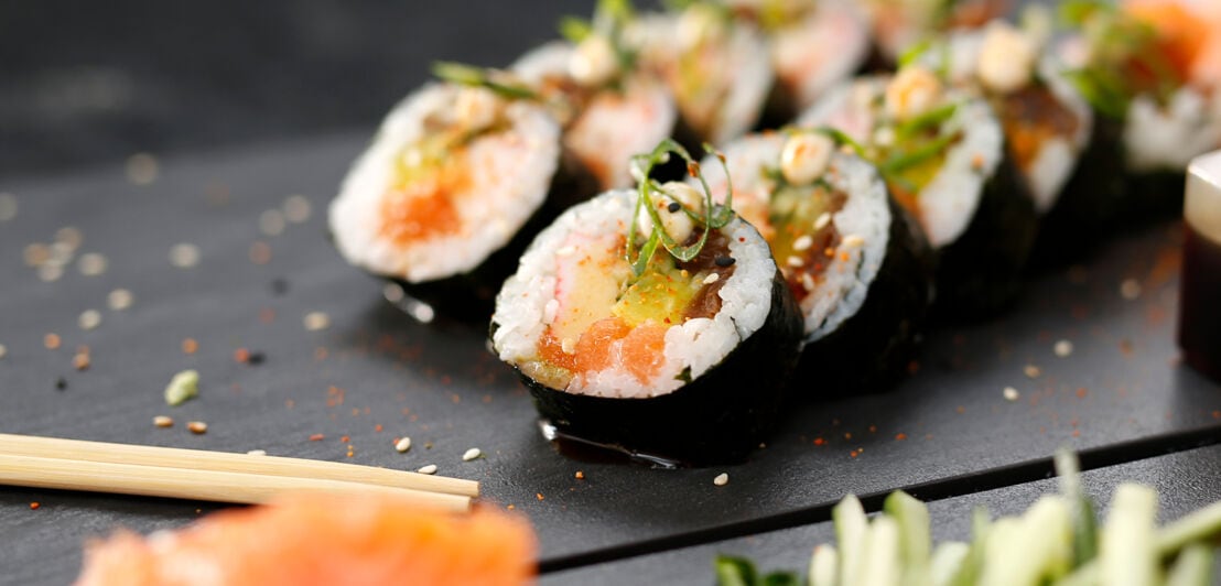 Nahaufnahme zweier geschnittener Sushirollen mit Fisch- und Gemüsefüllung auf schwarzem Untergrund