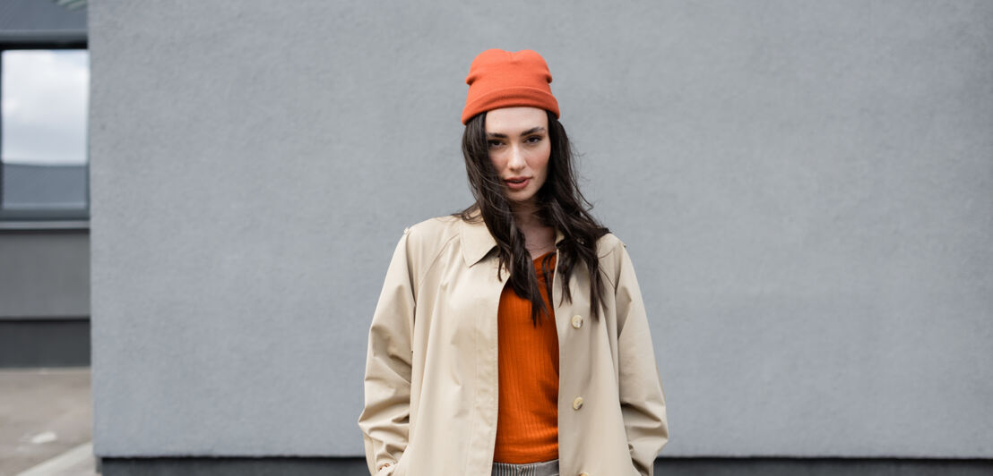 ine junge, moderne Frau, gekleidet mit beigem Trenchcoat, orangenem Pullover und orangener Mütze, posiert vor einer grauen Fassade mit Blick in die Kamera