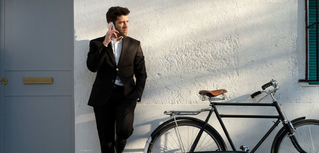 Ein junger Mann im Anzug lehnt telefonierend neben einem Fahrrad an einer Hauswand