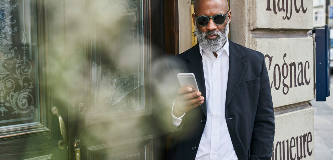 Ein Mann in Anzug und mit einer runden Sonnenbrille hat ein Handy in der Hand und lehnt an einer Hauswand
