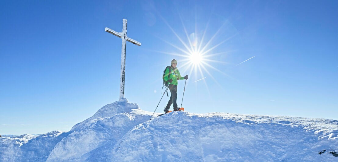 Ein Wanderer mit Schneeschuhen und Stöcken läuft neben einem Gipfelkreuz im Schnee