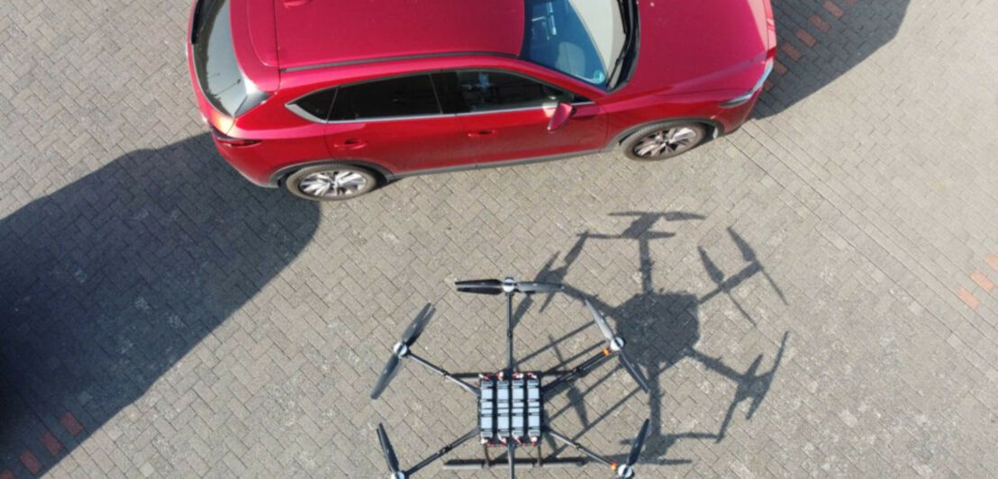 Eine große sechsarmige Drohne steht neben einem roten Kleinwagen und hat in etwa die gleiche Breite