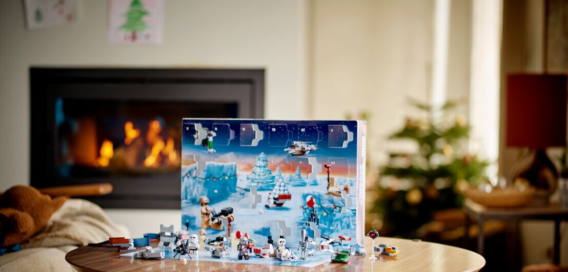 Ein geöffneter Adventskalender mit aufgebauten Lego-Spielfiguren steht auf einem Tisch in einem weihnachtlich geschmückten Wohnzimmer mit Kamin