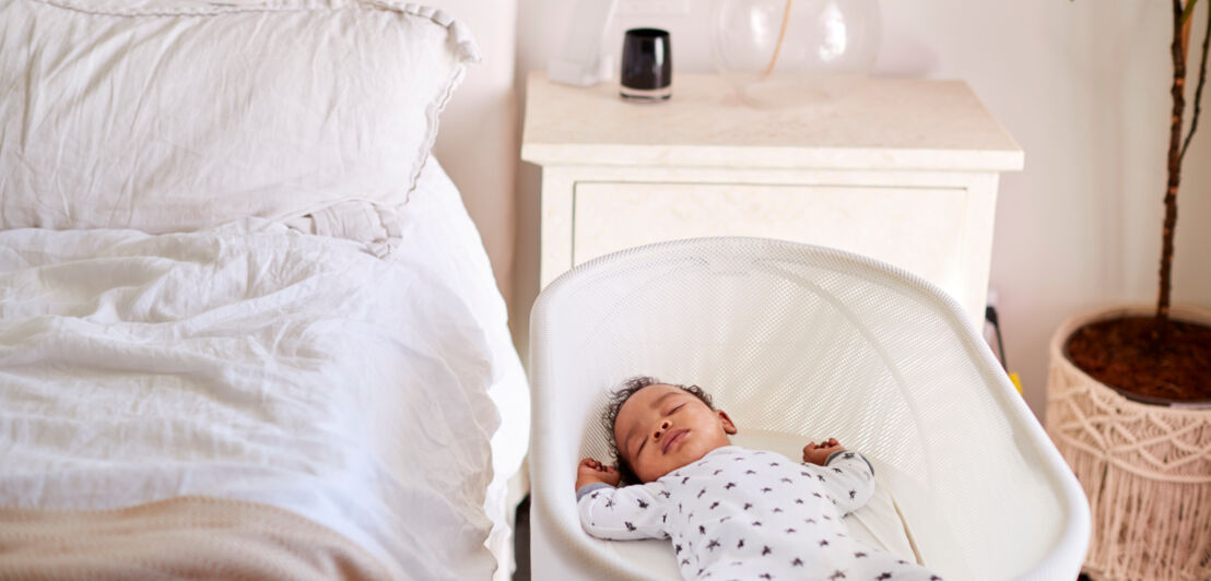 Ein Baby schläft in einem Wiegenbett, das neben dem Elternbett im Schlafzimmer steht