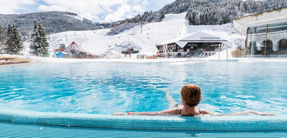 Eine Frau liegt in einem großen Pool, im Hintergrund sind verschneite Berge.