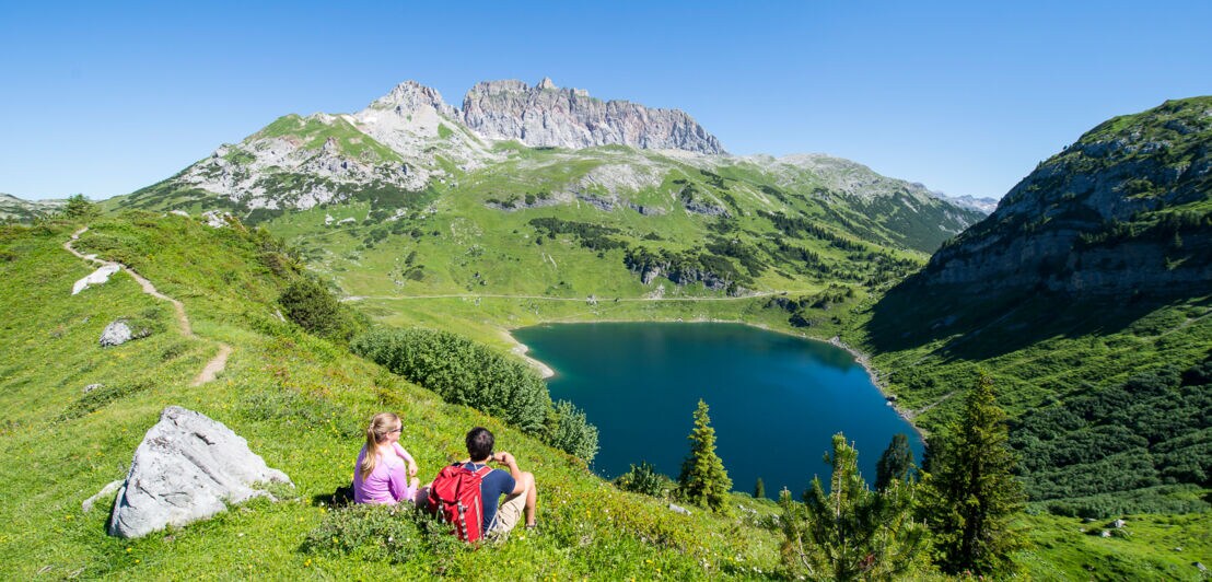 Zwei Wandernde sitzen auf einer Anhöhe vor einem See, im Hintergrund eine Bergkette