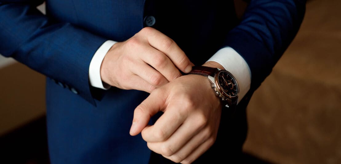 Nahaufnahme der Handgelenke eines Mannes in einem blauen Anzug, er berührt seine Armbanduhr