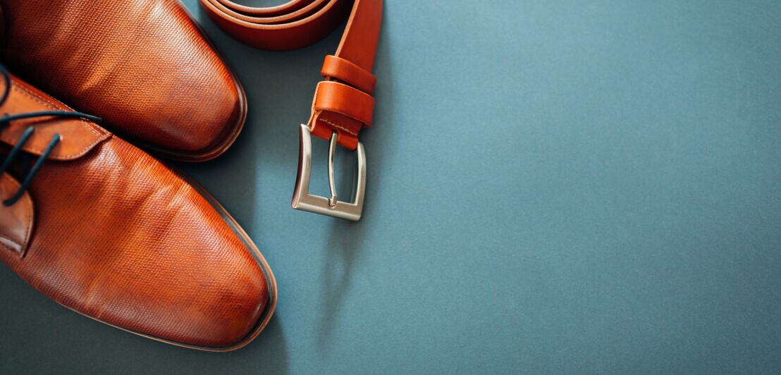 Ein Paar Lederschuhe und ein Ledergürtel in derselben Farbe vor türkisfarbenen Hintergrund