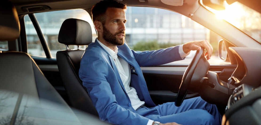 Ein Mann im Anzug sitzt in einem Auto, im Hintergrund Sonnenschein