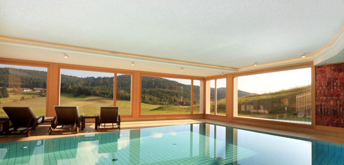 Minimalistischer Raum mit großem Innenpool, Liegestühlen und Panoramafenstern mit Blick in die bewaldete Landschaft
