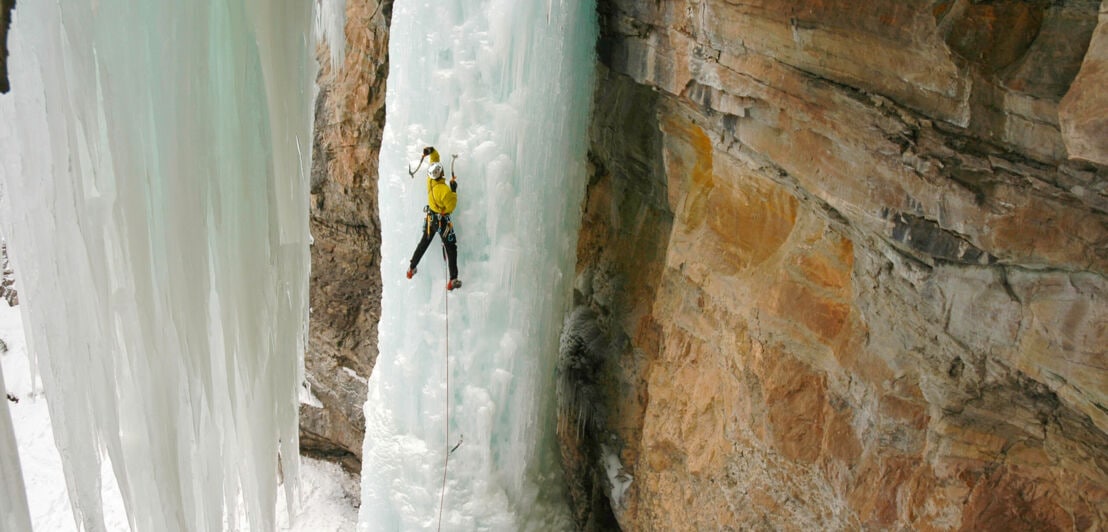 Eine Person klettert mit einem Seil gesichert einen gefrorenen Wasserfall hinauf.