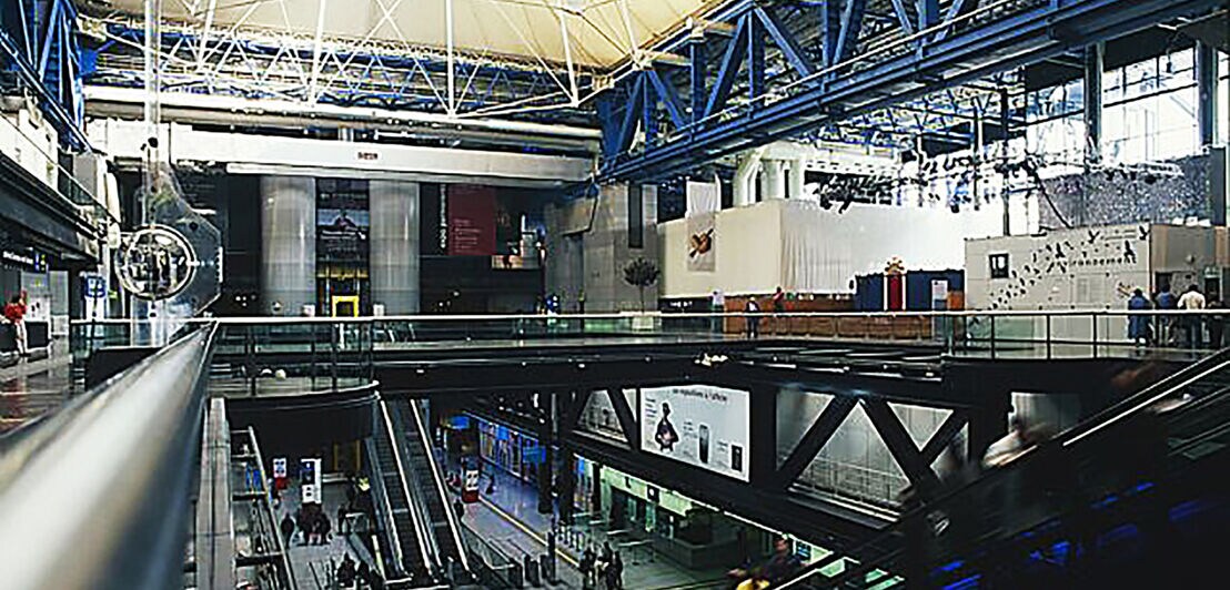 Innenaufnahme einer belebten Eingangshalle eines modernen, industriellen Gebäudes auf zwei Ebenen, die mit Rolltreppen verbunden sind