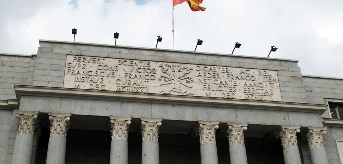 Mit Säulen geschmückter Eingangsbereich eines monumentalen Gebäudes mit Inschrift und wehender spanischer Flagge auf dem Dach