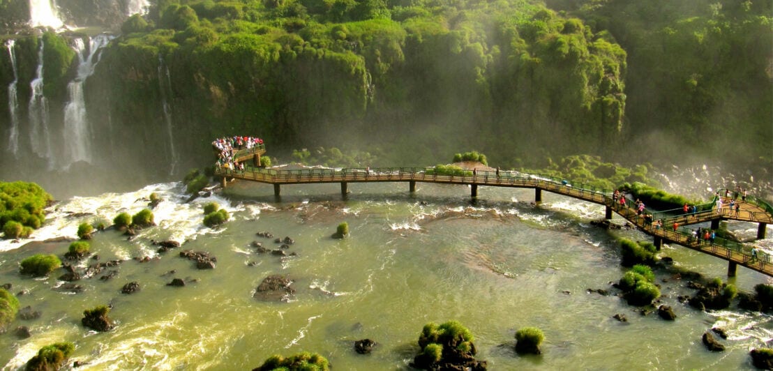 Panorama eines Naturparks mit Wasserfällen und Regenwald, in dem Besucher auf einem auf Stelzen gebauten Weg über das Wasser laufen