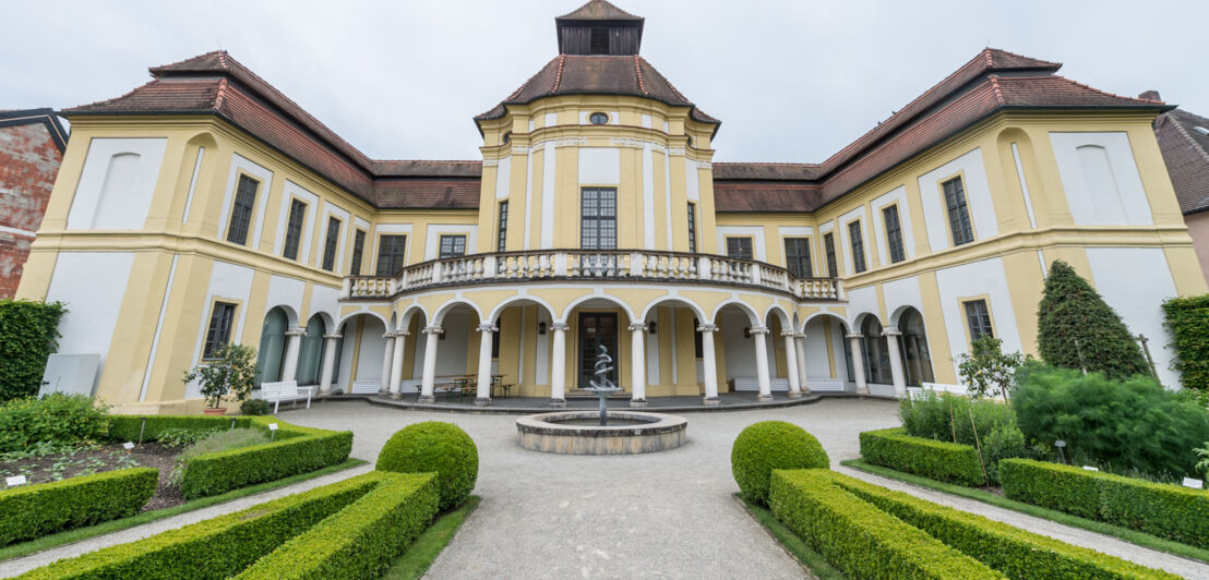 Das prächtige Gebäude des Deutschen Medizinhistorischen Museums mit weiß-gelber Fassade, davor Begrünung und ein Springbrunnen mit Äskulapstab