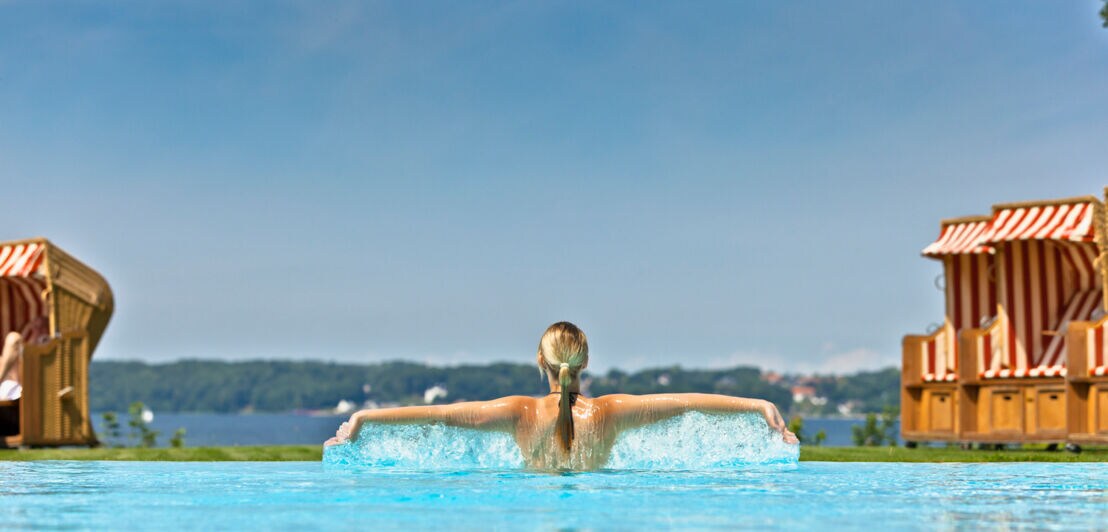 Rückansicht einer Frau im Pool mit ausgestreckten Armen, im Hintergrund Strandkörbe vor einer Meeresbucht