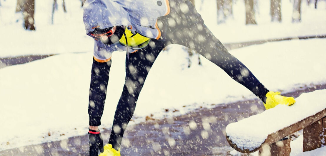 Ein sportlich gekleideter Mensch macht Stretchingübungen im Schnee.