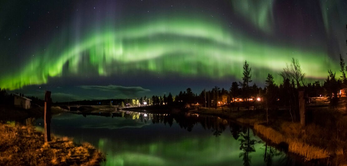 Grünes Polarlicht über Landschaftspanorama mit spiegelndem Wasser und einGrünes Polarlicht über Landschaftspanorama mit spiegelndem Wasser und einem beleuchteten Haus bei Nacht