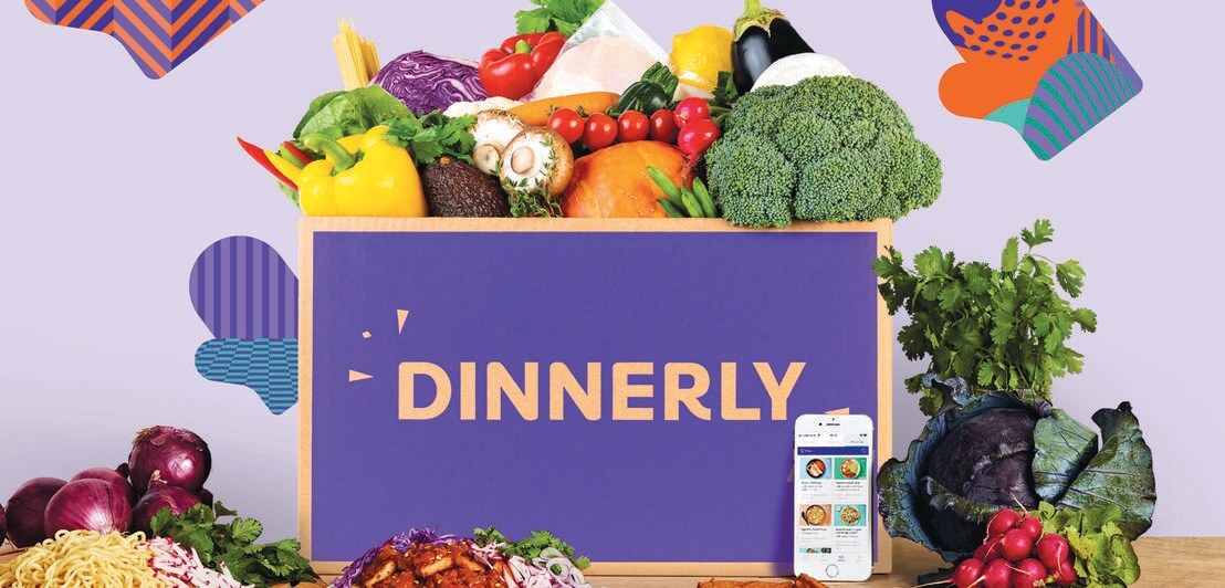 Eine lilafarbige Karton-Box mit Schriftzug steht auf einem Holztisch umgeben von frischen Lebensmitteln, davor ein Smartphone mit App und Gerichte auf Tellern