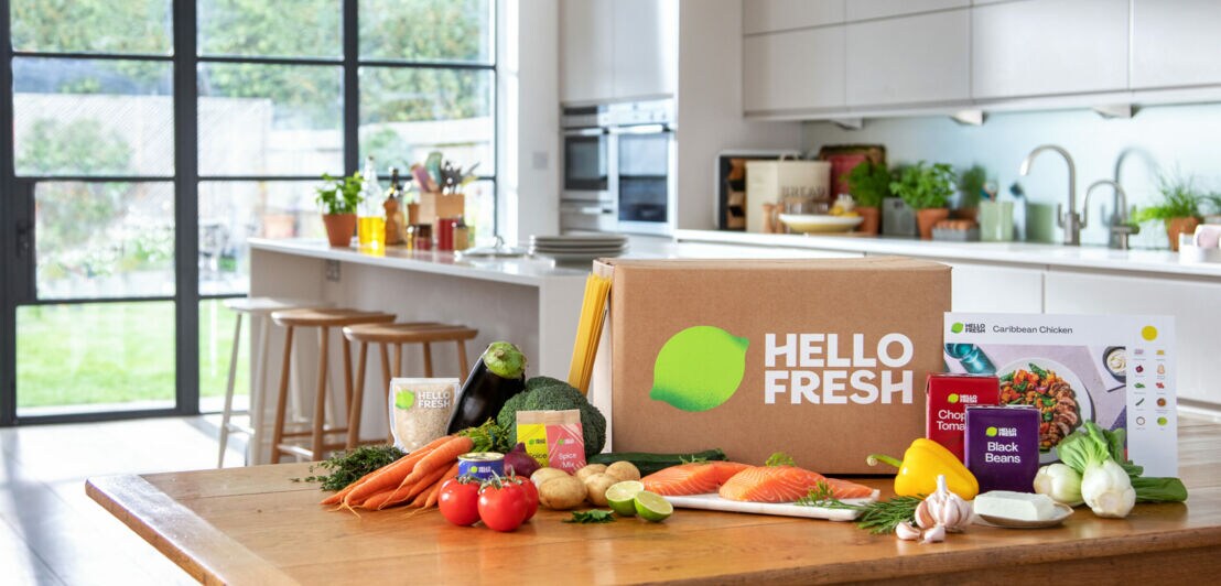 Eine Karton-Box mit Logo steht auf einem Holztisch umgeben von frischem Gemüse, Verpackungen und einer Rezeptkarte, im Hintergrund eine große, helle Küche mit Gartenblick