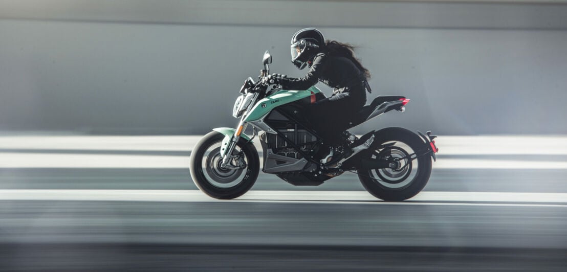 Ein mintgrünes Motorrad mit einer langhaarigen Fahrerin in schwarzer Kleidung