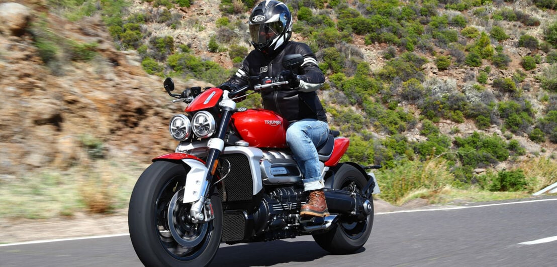 Ein rotes Motorrad fährt vor einer mediterran anmutenden Hügelkulisse durchs Bild.