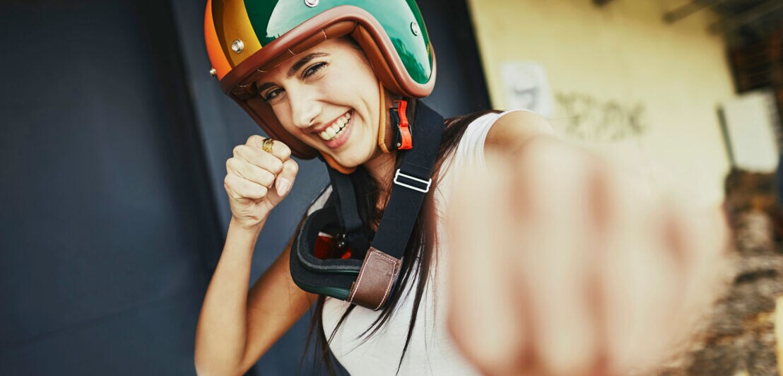 Eine junge, lachende Frau mit einem bunten Motorradhelm auf dem Kopf boxt spielerisch in Richtung Kamera