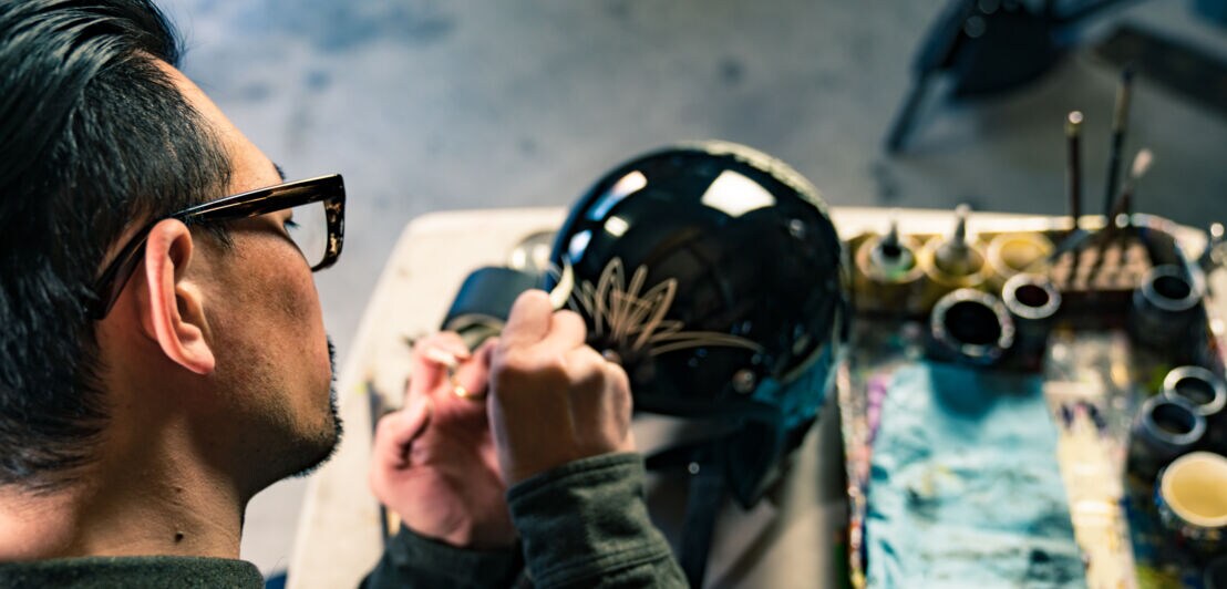 Blick über die Schulter eines Mannes, der einen schwarzen Motorradhelm mit goldenen Elementen bemalt, daneben Malutensilien in einem Atelier