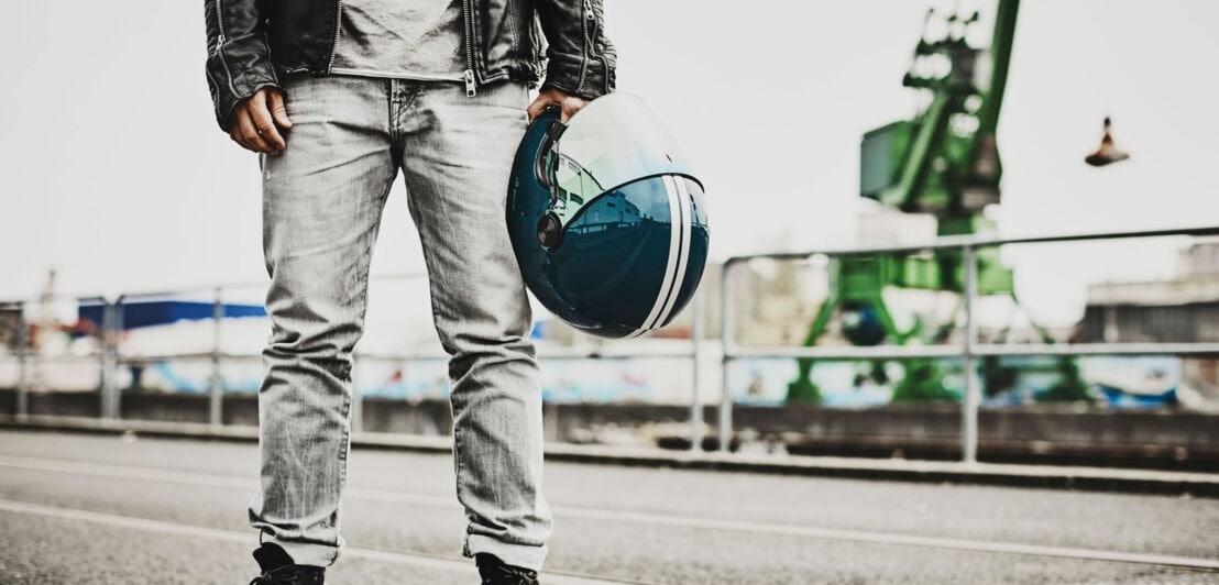 Unterkörper eines Mannes in lässiger Kleidung in einem Industriegebiet, der in einer Hand einen blauen Motorradhelm trägt