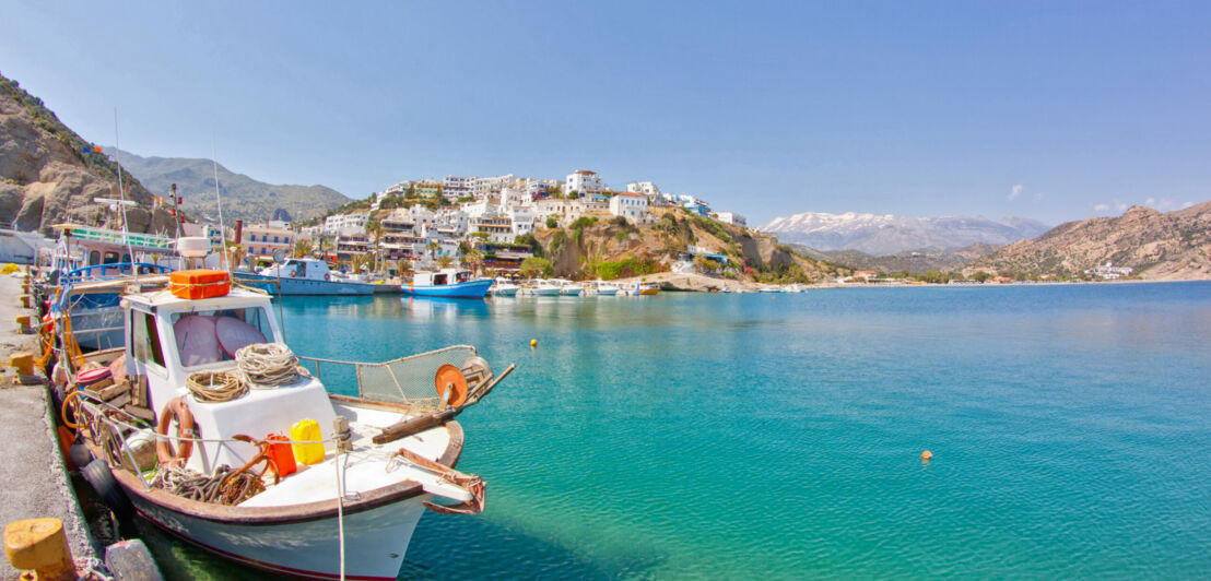 Kretischer Küstenort, der sich auf einen Hügel erstreckt, im Vordergrund ein Fischerboot am Anlegekai im türkisblauen Wasser