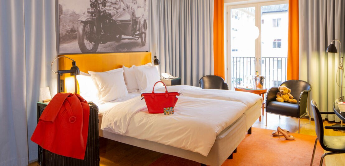 Hotelzimmer. Im Mittelpunkt ein Bett, daneben Koffer, Stühle, Vorhänge