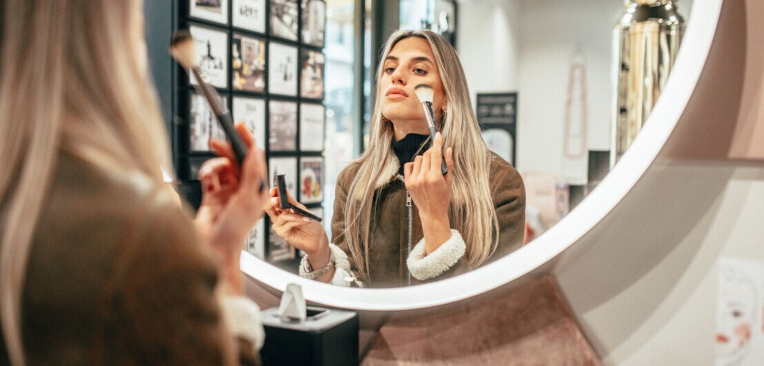 Eine Frau steht vor einem großen runden Spiegel und pudert sich das Gesicht